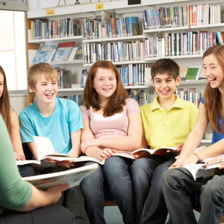 Lärare och sex barn i nedre tonåren med böcker i händerna pratar i bibliotek