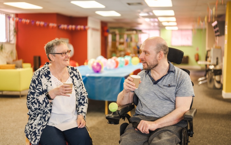 Two elderly people drinking from paper mugs. The man is in a wheelchair. Baloons in the background.