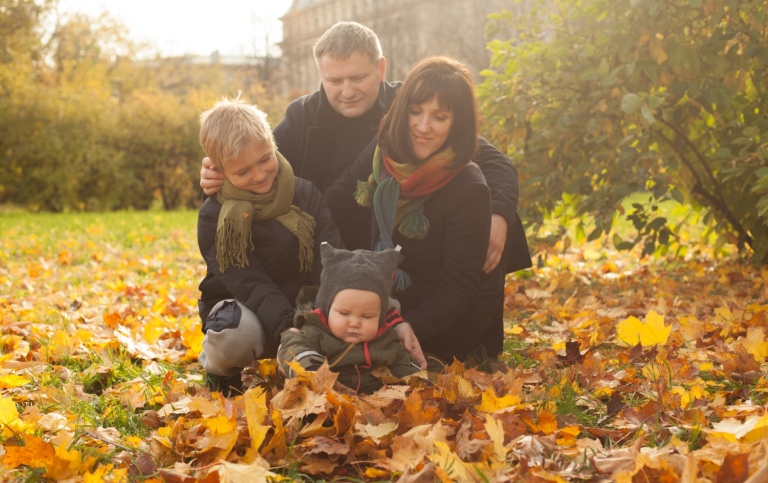 Mother, father and older brother looking at baby playing with autumn leaves.