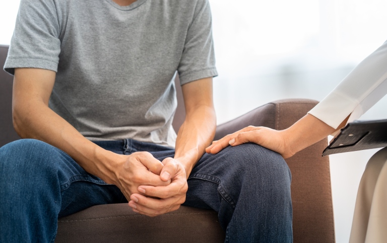 En person sitter framåtlutad med händerna knäppta, medan en annan lägger en hand på personens knä.