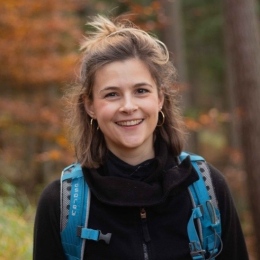 The portrait photo shows the researcher Ronja Kraus in a forest wearing a black jacket and blue backpack and smiling to the camera. Profile picture of Ronja Kraus