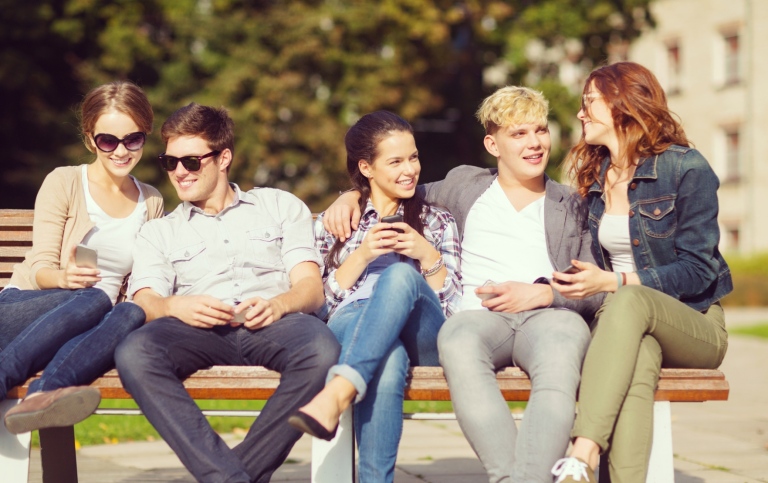 High school students – three girls and two boys – sitting outside on a bench, talking, smiling and l