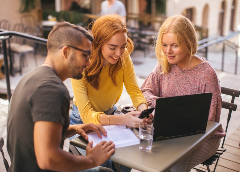 Tre studenter sitter vid cafébord utomhus och tittar på mobil och laptop tillsammans.