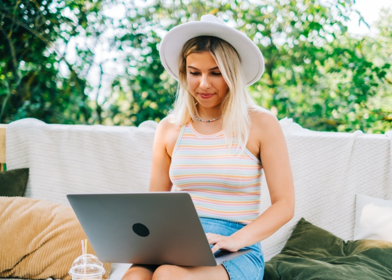 Ung sommarklädd kvinna sitter ute och skriver på laptop i knät.
