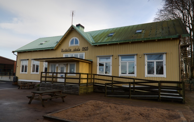 Gul trähusbyggnad med grönt plåttak. Text på huset: Kvibille skola 1905.