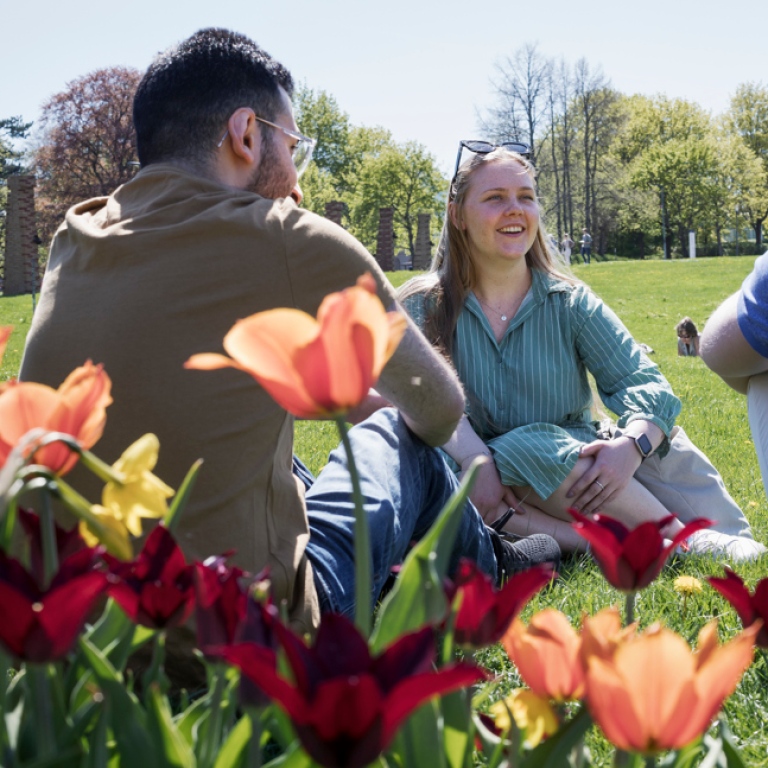 Studenter sitter i gröngräset, blommor i förgrunden.