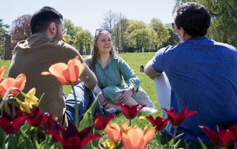 Studenter sitter i gröngräset, blommor i förgrunden.