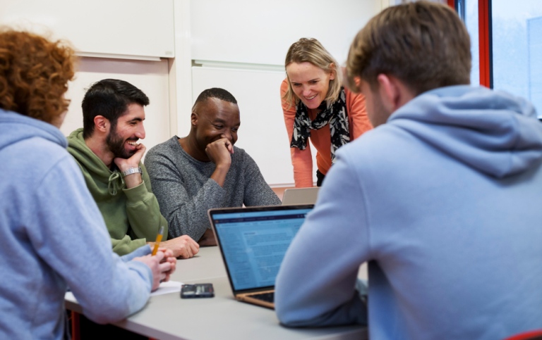 Students sitting around a table with a laptop, and teacher standig talking to them.