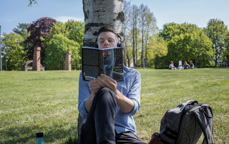 A male student is sitting under a tree, reading a book.