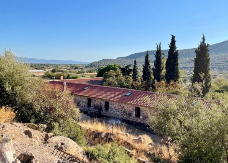 Low brick buildings with red roof, in a greek landscape with mountains, cypress trees, dry grass.