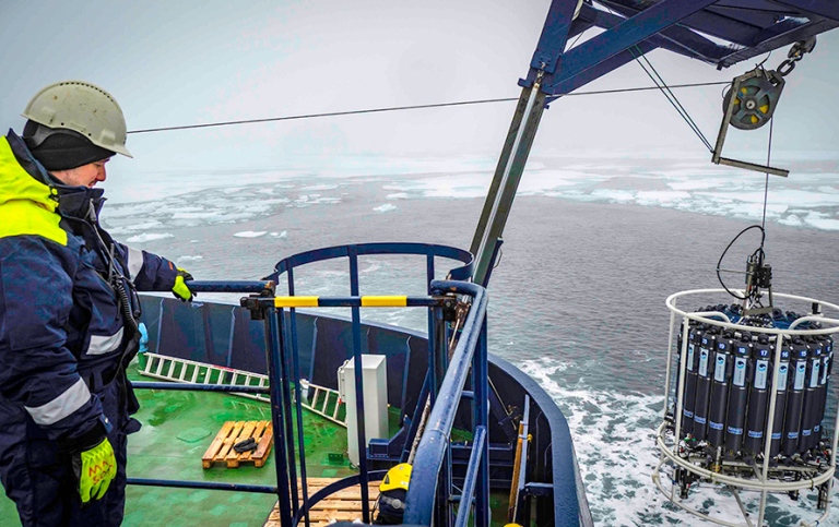 A researcher in helmet and outdoor clothes on icebreaker Oden looks at water sample "bottles".