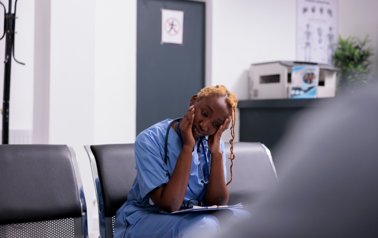 Tired healthcare worker in blue scrubs sitting in a waiting area with hands on face.