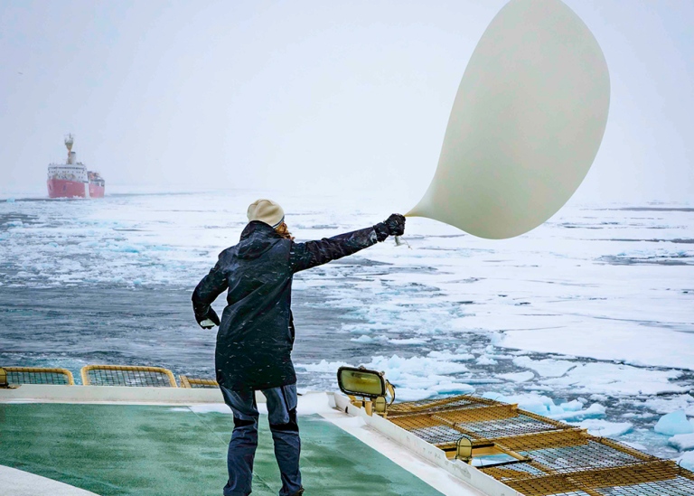 Student on deck struggling with big balloon in the wind. Icebreaker St Luis in the background.