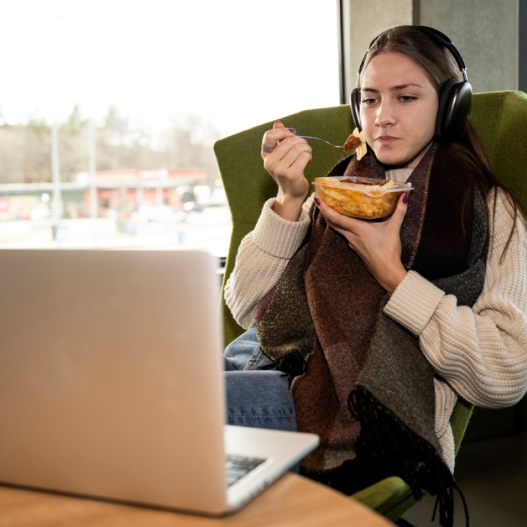Kvinnlig med hörlurar student sitter i läsfåtölj, äter matlåda och tittar på laptop. 
