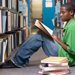 En ung student i grön t-shirt sitter på golvet mellan bokhyllor i ett bibliotek och läser en bok. 