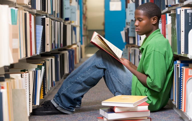 En ung student i grön t-shirt sitter på golvet mellan bokhyllor i ett bibliotek och läser en bok. 