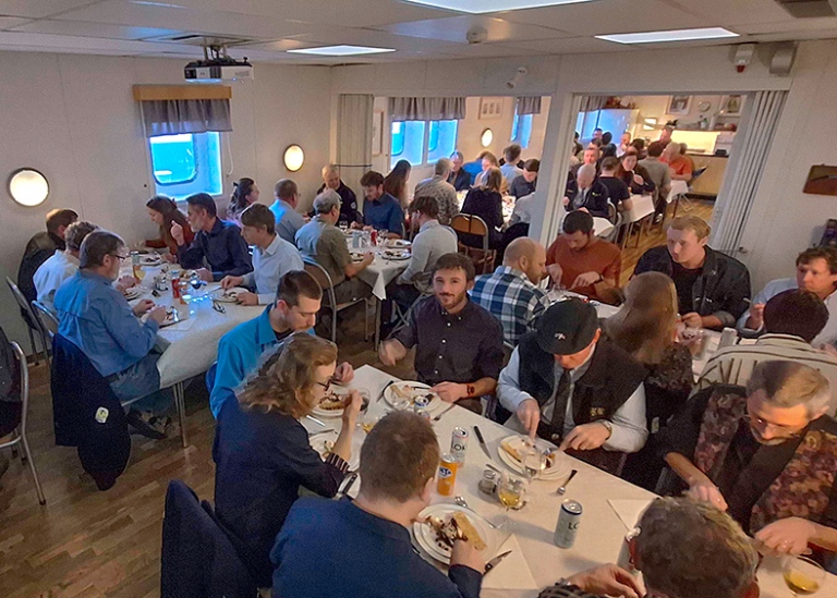 Up to ten tables with 6–8 people at each table, with white tableclotns, having dinner.