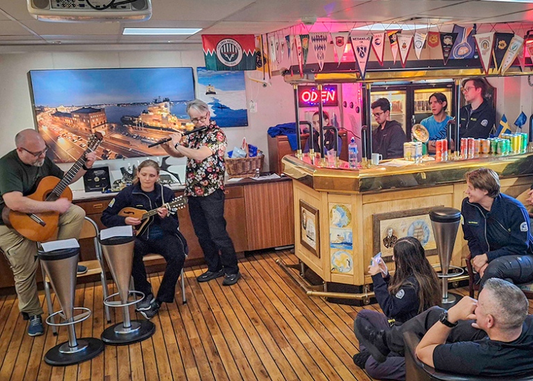 Tre personer spelar instrument, en bar och sittande publik.