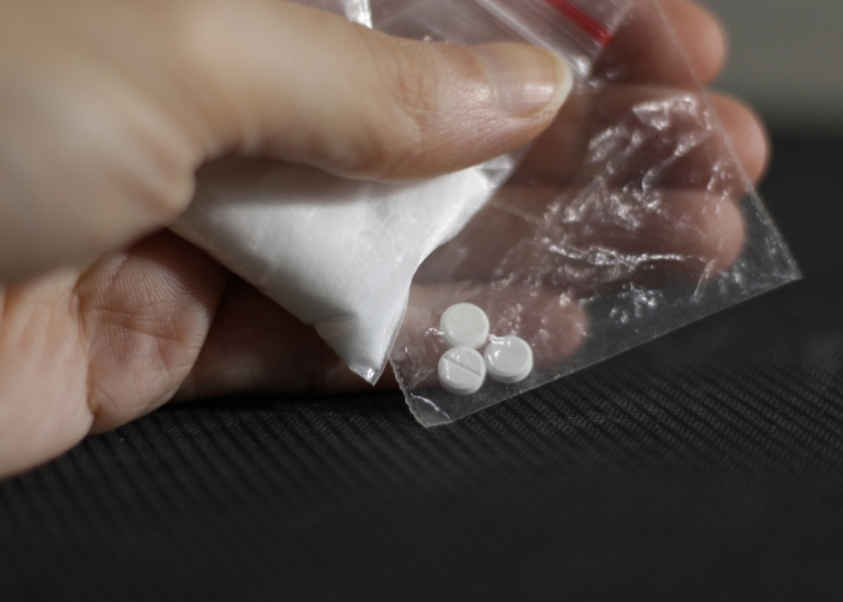 Close up of a hand holding two transparent bags, one with cocaine and one with pills.