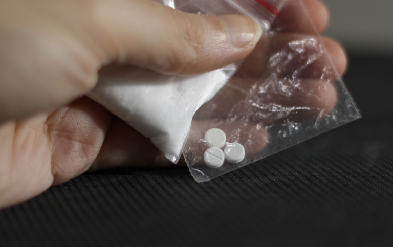 Close up of a hand holding two transparent bags, one with cocaine and one with pills. 