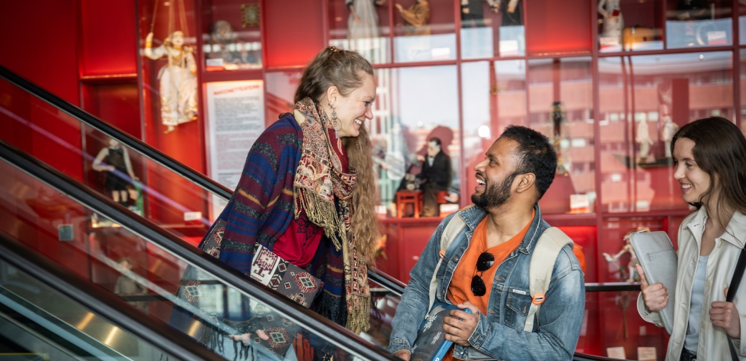 Studenter samtalar och skrattar i en byggnad med röd inredning. Två av dem står på en rulltrappa och