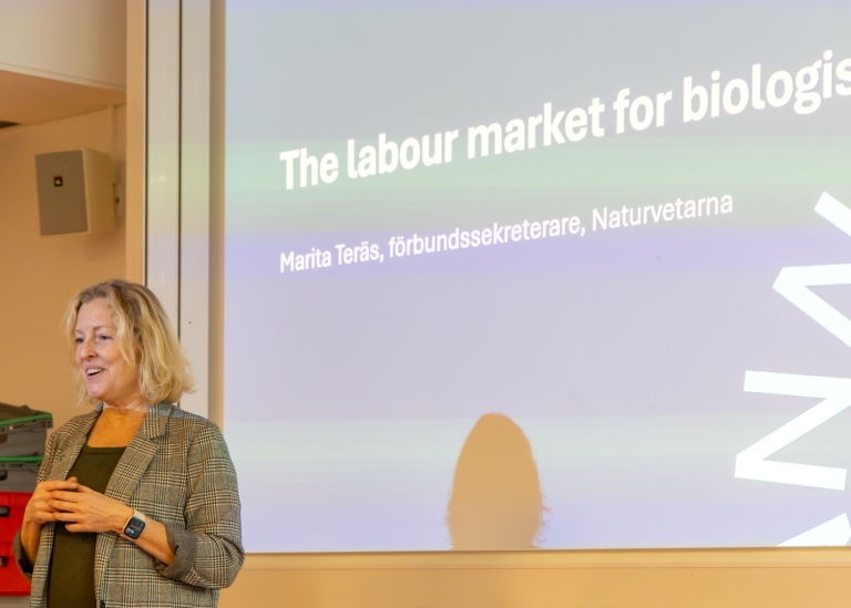 Maria is blond and stands in front of a presentation of Naturvetarna