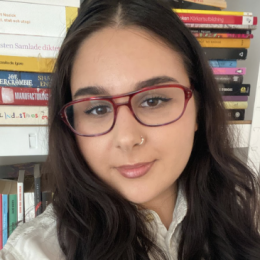 A woman with light tan skin, dark brown eyes and long dark brown hair smiles in front of a bookshelf
