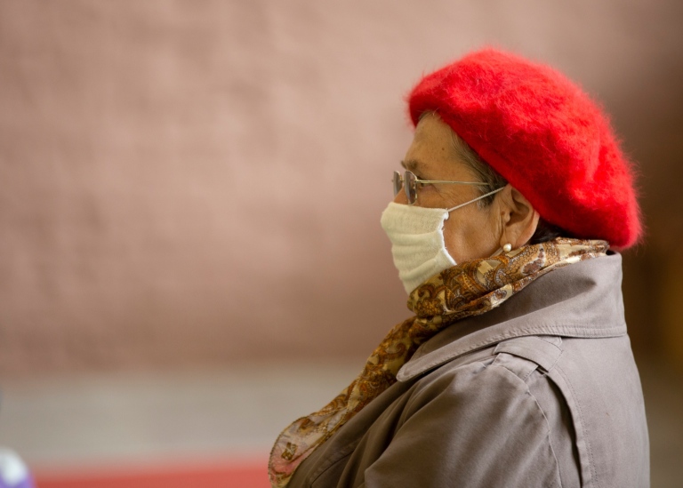 Elderly woman sitting wearing a beret and a surgical mask.