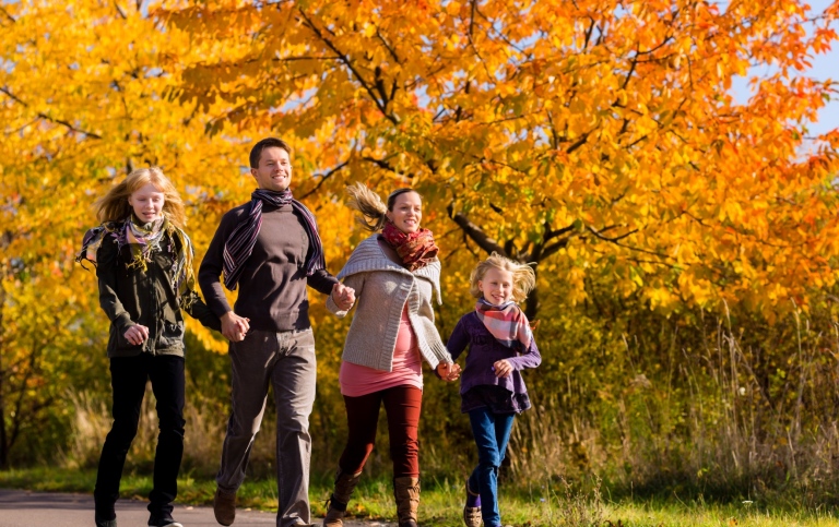 Två vuxna och två barn småspringer på en stig i en park på hösten.