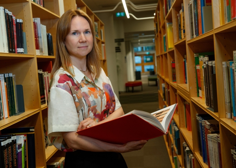 Kvinna står i ett bibliotek och håller upp en bok. 