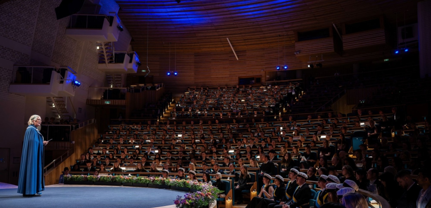 A person stands on stage in Aula Magna in front of a large audience during the master's ceremony.