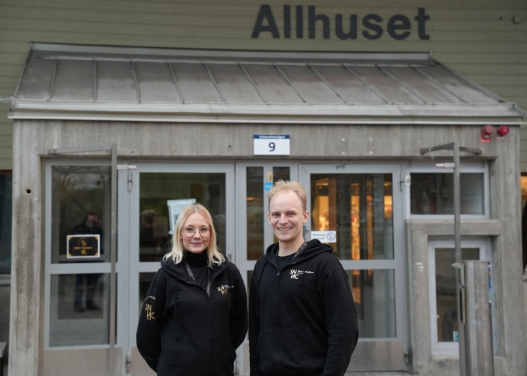 Olivae Odenskiöld och Miro Jääskeläinen i arbetskläder framför entrén till Allhuset